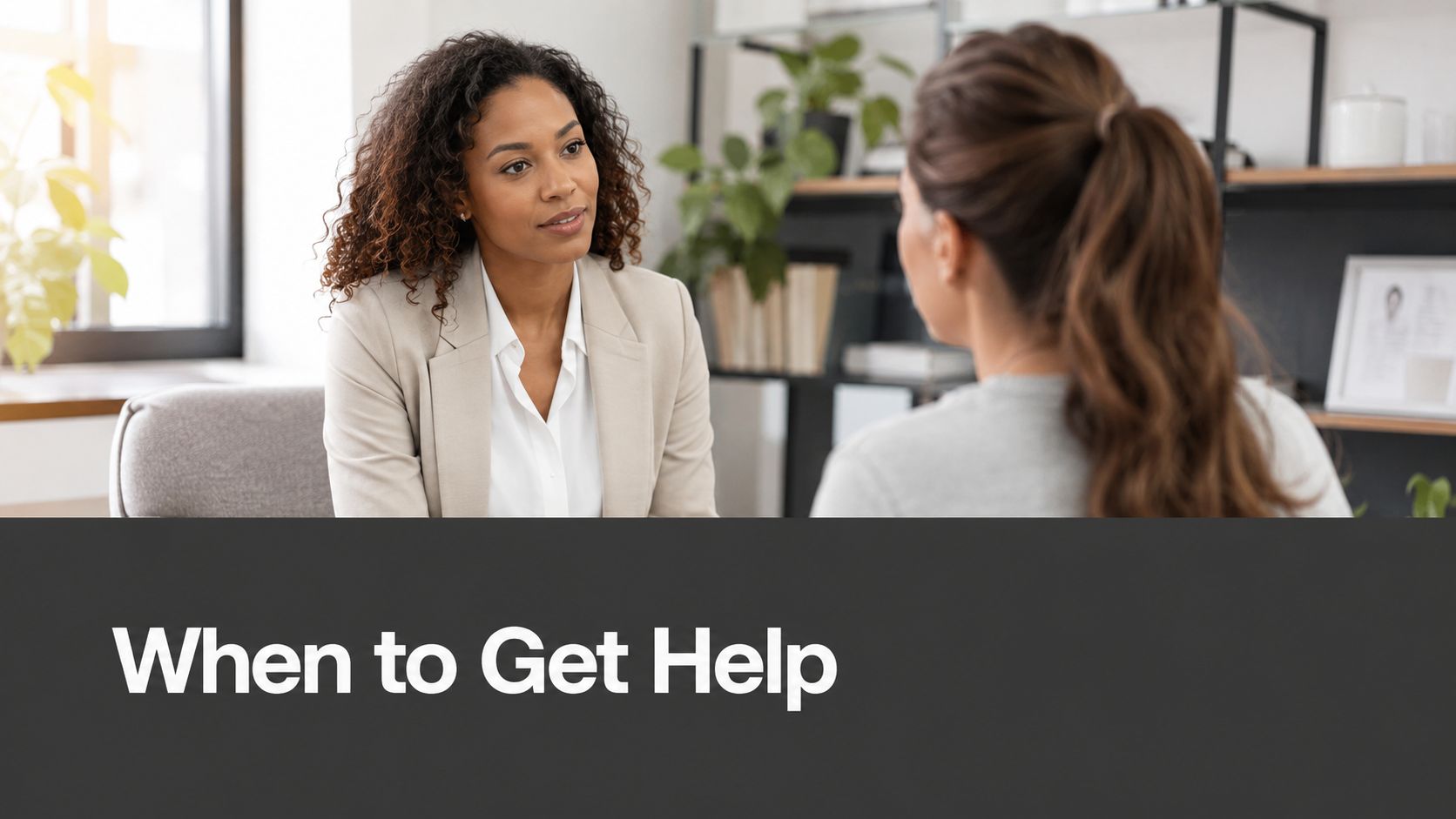 A professional woman in a beige blazer listens attentively to her client in an office setting.